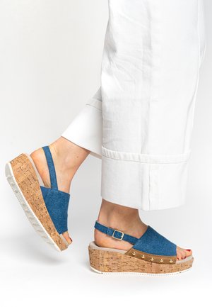 Denim wedge sandals with cork sole, blue upper, adjustable buckle, and gold accent studs. Worn with white wide-leg pants.