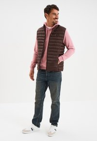 Lächelnder Mann steht und schaut nach rechts, trägt eine braune Steppweste über einem rosa Hoodie, blaue Jeans und weiße Sneakers mit marineblauen Streifen.