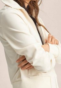 Sudadera blanca con cremallera hecha de un tejido suave, de corte relajado, con puños acanalados y bolsillos laterales. Presenta un cuello alto y una textura suave.