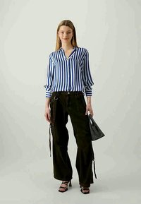 Ung kvinna klädd i en blå och vit vertikalt randig skjorta, svarta vida byxor med remmar, svarta klackskor och håller en liten svart handväska.