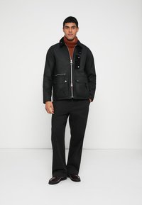 Black quilted jacket with a zip front, multiple pockets, and a contrast collar; paired with dark pants and brown shoes against a plain background.