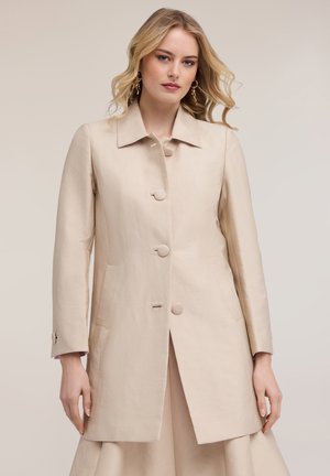 Vaaleahiuksinen nainen, joka pukeutunut pitkään beigenväriseen nappilistalliseen takkiin, jossa on kaulus, ja kultaisiin rengaskorvakoruihin, yksinkertaista vaaleaa taustaa vasten.