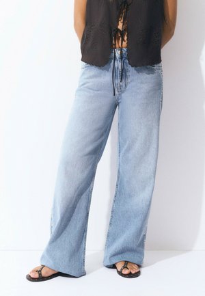 Light blue wide-leg jeans made of denim, featuring a high waist and side button accents. Paired with a black embroidered top and flat sandals.