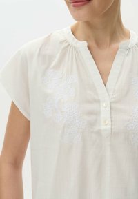 Světle béžová blůza s krátkými rukávy, která má bílé květinové vyšívání na hrudi a zapínání na knoflíky s kulatým límcem.