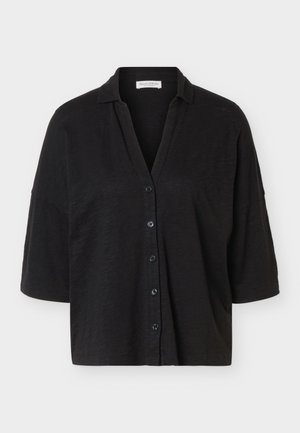 Crna košulja širokog kroja s gumbima, širokim rukavima srednje dužine i malim ovratnikom, prikazana na jednobojnoj svijetloj pozadini.