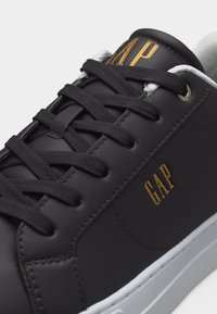 Baskets en cuir noir avec lacets noirs, semelle blanche et logo "GAP" doré sur la languette et le côté.