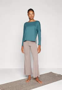 Bluza verde-albastră cu mâneci lungi și tăieturi laterale, asortată cu pantaloni beige evazați de yoga. Material neted și croială lejeră, potrivită pentru activități sportive.