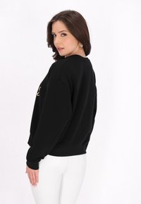 Felpa nera con scollo rotondo e maniche lunghe, caratterizzata da una scritta dorata sul davanti, indossata con pantaloni bianchi attillati.
