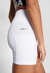 Shorts bianchi a vita alta con una texture liscia, caratterizzati da dettagli a pannello e da un piccolo logo "NEBBIA" sul retro. Design aderente.