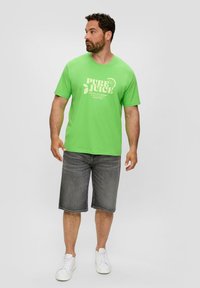 Helles grünes Baumwoll-T-Shirt mit dem Aufdruck "PURE JUICE" in Weiß, kombiniert mit grauen Jeansshorts und weißen Sneakers.