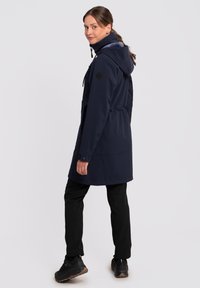 Parka imperméable bleu marine avec capuche, taille élastique, deux poches avant et tissu texturé. Un pantalon noir et des chaussures complètent l'ensemble.