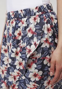 Close-up of a floral skirt with white and red hibiscus flowers and blue leaves, featuring an elastic waistband and a side pocket, worn by a person.