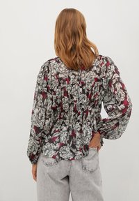 Blusa floreale nera con motivo bianco e rosso, tessuto leggero e trasparente, maniche a palloncino, dettagli arricciati sulla schiena, abbinata a jeans grigi chiari.