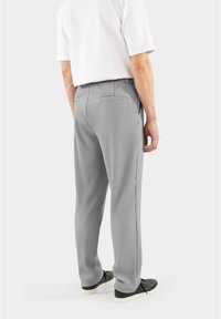 Graue, plissierte Hose mit glattem Stoff, ausgestattet mit seitlichen Taschen und einer Gesäßtasche, kombiniert mit schwarzen Sneakers und einem weißen Hemd.
