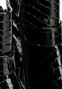 Primer plano de botas de charol negro brillante con textura de cocodrilo y cremallera visible en el costado.