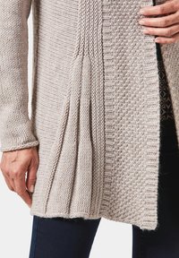 Hellbeige gestrickte Strickjacke mit strukturierten Mustern, offenem Vorderteil und Falten am Saum. Weiches, dickes Garn und gerippte Bündchen.