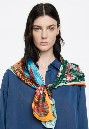 Zijde sjaal met kleurrijke bloemen- en veerpatronen in oranje, blauw en groen, geknoopt om de nek, gedragen over een blauwe overhemd met knopen.