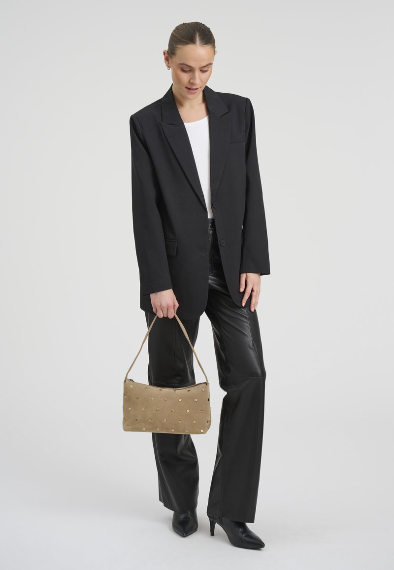 Femme portant un blazer noir surdimensionné, un pantalon en cuir noir, des bottes à talons noires, un haut blanc, tenant un sac à main beige à clous dorés.