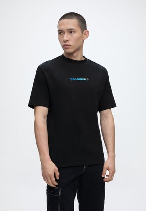 Mann mit rasiertem Kopf, der ein schwarzes Karl Lagerfeld T-Shirt und schwarze Hose trägt, steht vor einem schlichten weißen Hintergrund.