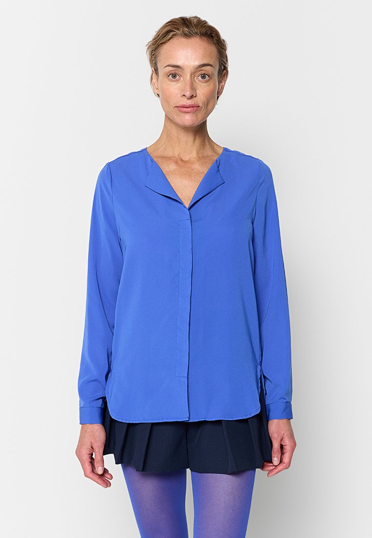 Blaue Langarmbluse mit V-Ausschnitt, Vorderblende und leichter Raffung an den Schultern, kombiniert mit einem dunklen Rock und lila Strumpfhosen.