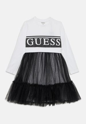 Langärmliges Kleid mit einem weißen Oberteil, das mit einem schwarzen "GUESS"-Logo versehen ist, und einem Rock aus schwarzem Tüll mit einem gerafften Saum.