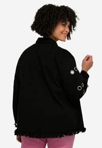 Zwarte corduroy jas met franjes aan de onderkant, oogjes details op de mouwen en een kraag. Model van achteren getoond, draagt roze broek.