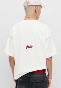 Hvid bomulds t-shirt med korte ærmer, der har et burgunder broderet logo på ryggen. Stylet med denim jeans og et rødt bælte.