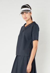 Top blu navy a maniche corte con colletto a punta e vestibilità rilassata, abbinato a una gonna coordinata. Visiera trasparente con dettagli bianchi indossata come copricapo.