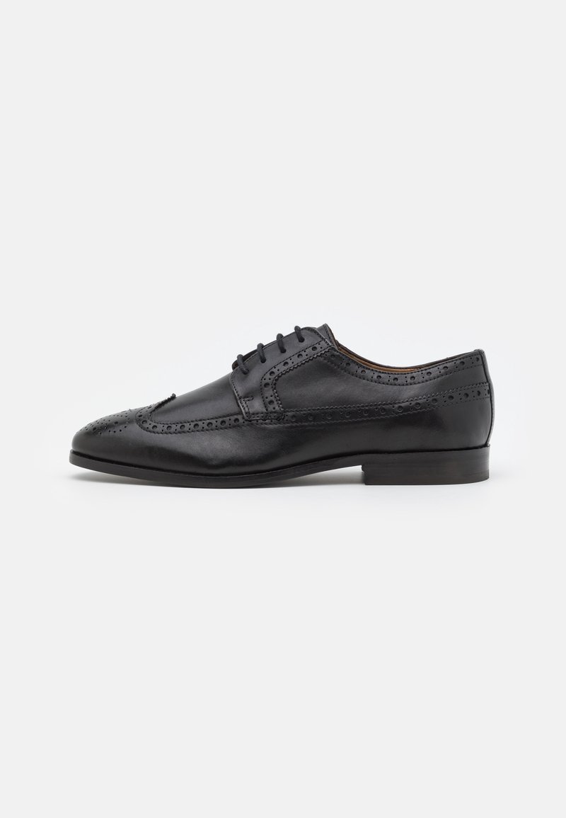 Chaussure de ville en cuir noir avec des détails de brogue, à lacets, à bout arrondi et à talon bas empilé. Texture lisse avec finition polie.