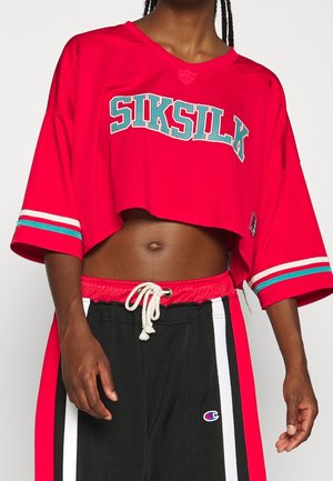 Červený zkrácený svetr s nápisem "SIKSILK" v barvě teal, krátké rukávy a bílé a teal pruhované detaily na rukávech. Černé tepláky s bílými proužky.