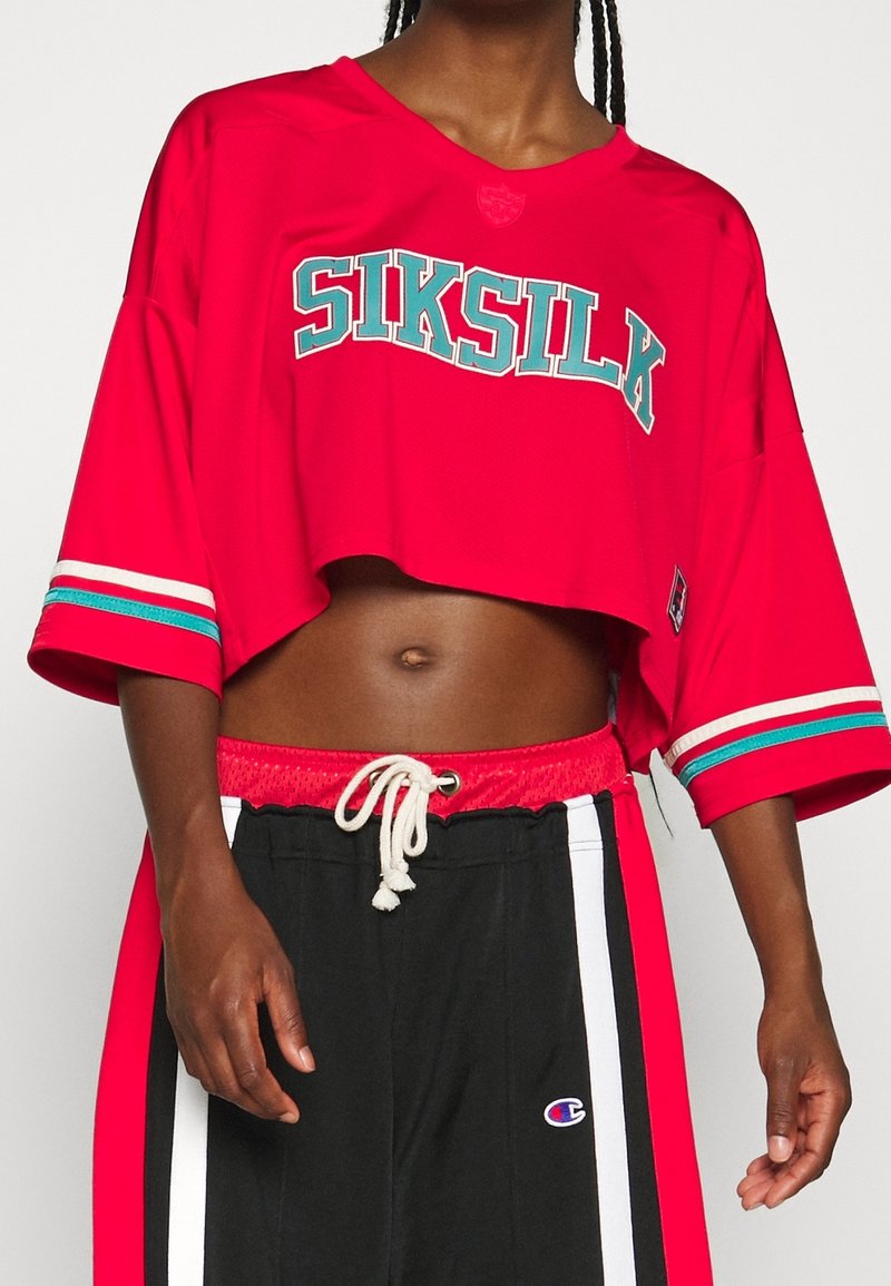 Rotes, kurzes Jersey mit "SIKSILK"-Schriftzug in Türkis, ausgestattet mit kurzen Ärmeln und weißen sowie türkisen Streifenakzenten an den Ärmeln. Schwarze Jogginghosen mit weißen Streifen.