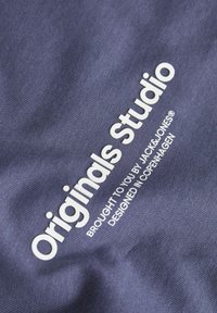 Jūrų mėlynumo audinys su sklandžiu paviršiumi, ant kurio balta spalva parašyta: "Originals Studio" ir papildomas tekstas apačioje, nurodantis prekės ženklą ir dizaino kilmę.