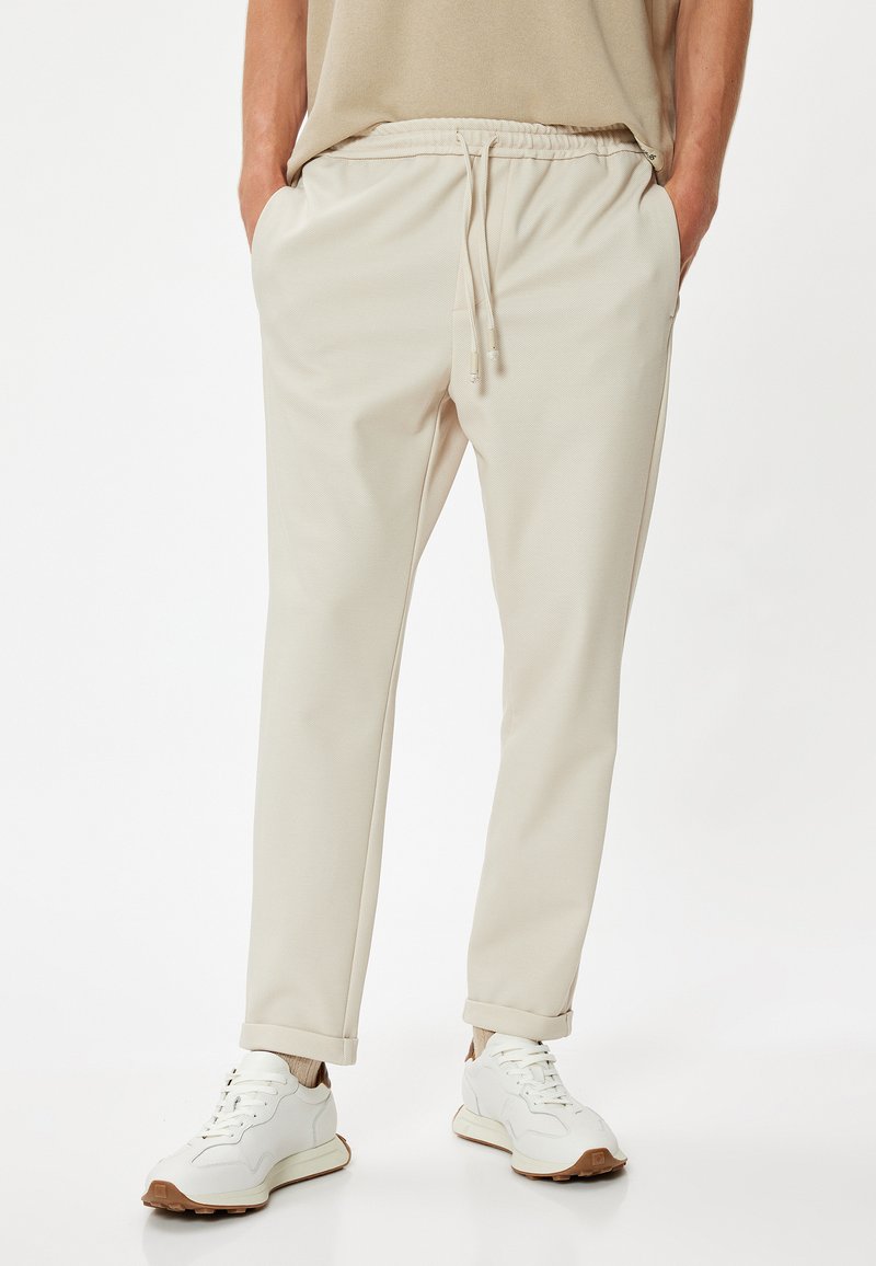 Pantalones jogger de color beige claro con un material texturizado, cintura elástica con cordón, bolsillos laterales y dobladillos ajustados. Combinados con zapatillas blancas.