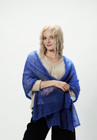 Niebieski, półprzezroczysty sweter w formie narzutki zarzucony na jasnobeżową bluzkę, z luźnymi włóknami i falistym wykończeniem, noszony w swobodny sposób.