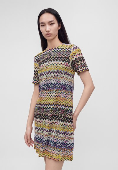 Femme aux longs cheveux noirs portant une robe à manches courtes avec des motifs en zigzag multicolores, debout devant un fond uni.