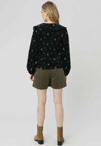 Chemisier noir avec des motifs floraux, manches bouffantes et col volanté, associé à un short en velours côtelé vert olive et des bottines beige.