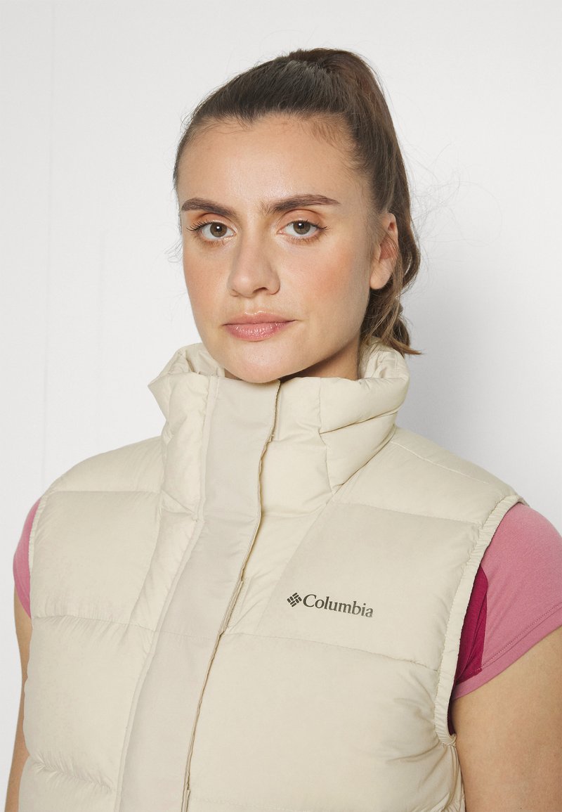 Beige Columbia-Steppweste mit hohem Kragen, ausgestattet mit einem Frontreißverschluss und gestepptem Design. Logo auf der linken Brustseite angebracht.