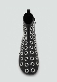 Botín negro de cuero con grandes ojales metálicos dispuestos en un patrón, con cremallera lateral y un tirador en forma de bucle.