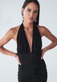 Crna halter haljina s dubokim V-izrezom i nabranim detaljem u struku, s elegantnom siluetom i glatkim materijalom.