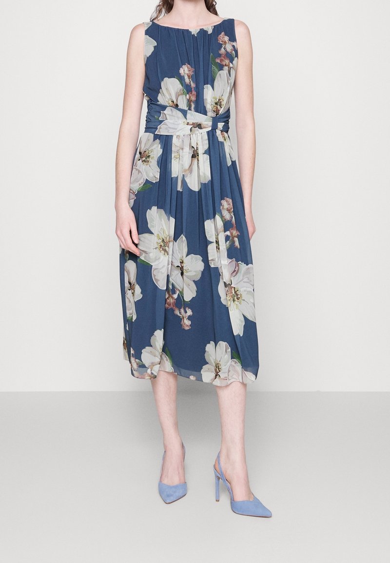 Femme portant une robe midi bleu sans manches avec de grands motifs floraux blancs et une ceinture assortie, associée à des chaussures à talons hauts bleu clair.