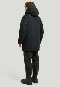 Svart isolerad jacka med huva, i kombination med vattentåliga byxor och svarta stövlar. Slät textur, minimalistisk design, praktisk passform.