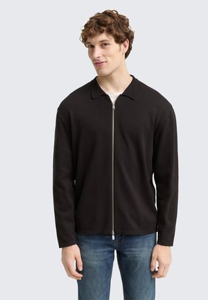 Black zip-up jacket with a point collar, made from a smooth fabric. Features long sleeves and a straight hem, worn over a white shirt.