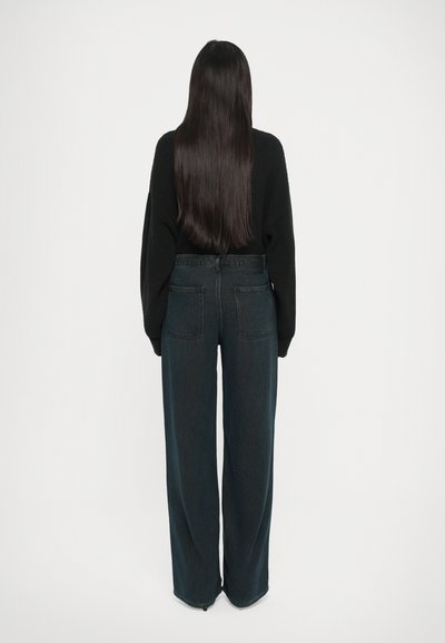 Jeans de corte ancho en denim oscuro con una cintura alta, bolsillos traseros y un suéter negro texturizado. El cabello cae recto hasta la mitad de la espalda.