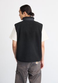 Melnas fleece veste ar augstu apkakli, teksturētu audumu un aizmugurējām šuvju detaļām, valkāta virs gaiša t-kreklu un saskaņota ar pelēkiem cargo biksēm.