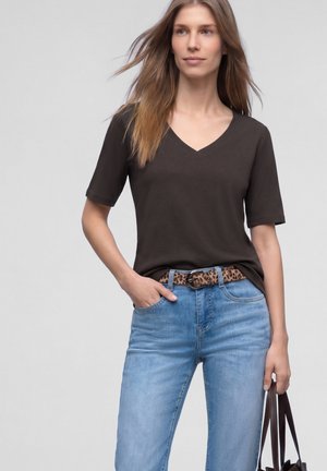 Ung kvinde iført en sort V-hals trøje, lyseblå jeans med et leopardprint bælte, der holder en mørkebrun håndtaske mod en grå baggrund.