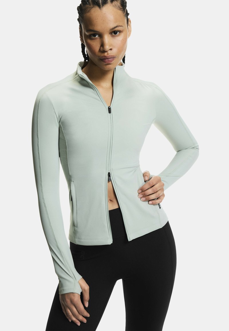 Kvinde med flettet hår iført lys grå lynlåstrøje med tommelfingerhuller og sorte leggings, poserer med den ene hånd på hoften mod hvid baggrund.