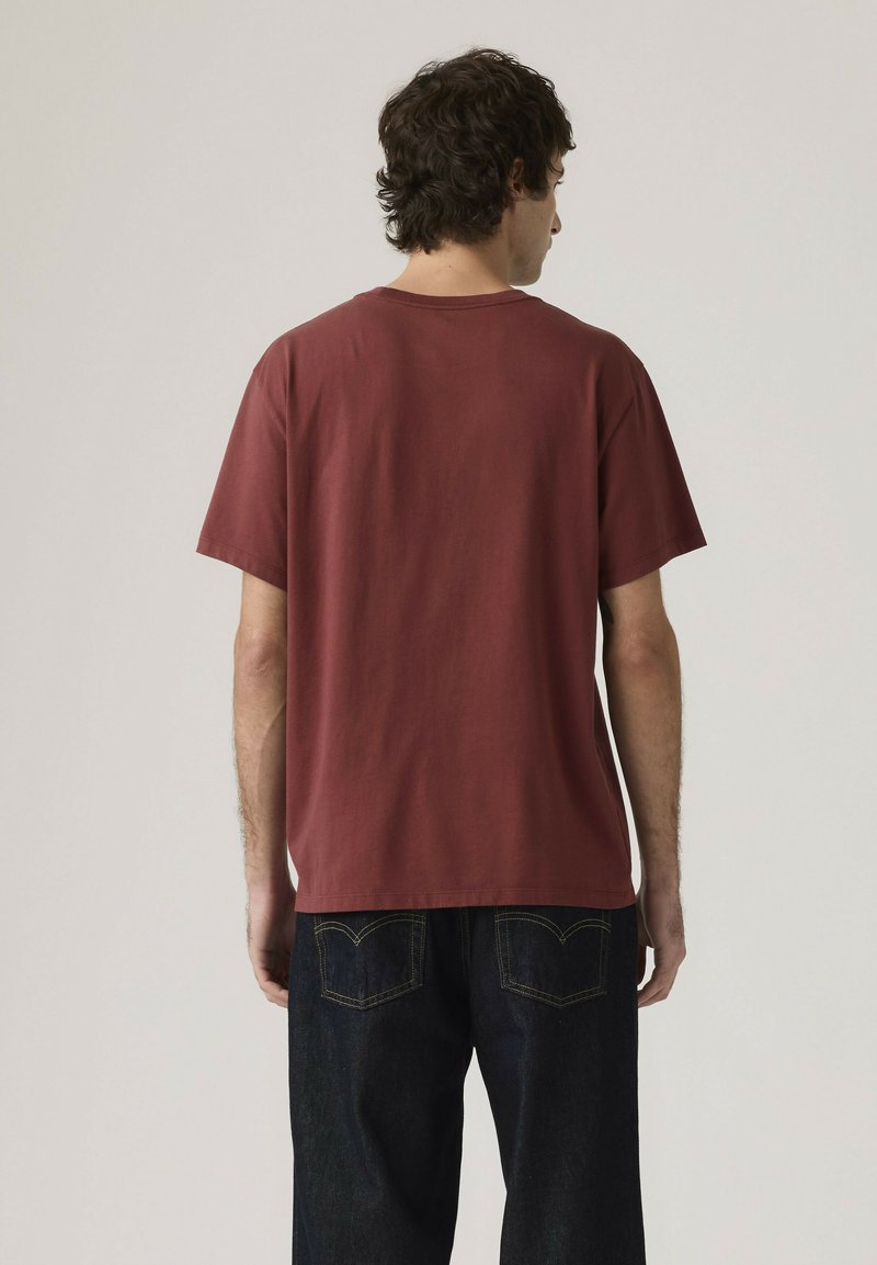 Uomo in piedi di spalle che indossa una maglietta a maniche corte color marsala semplice e jeans blu scuro su uno sfondo neutro.