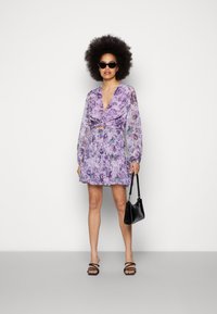 Vestido com estampado floral em tons de roxo, mangas longas transparentes, decote em V, detalhe de laço na cintura, saia rodada; combinado com acessórios pretos.