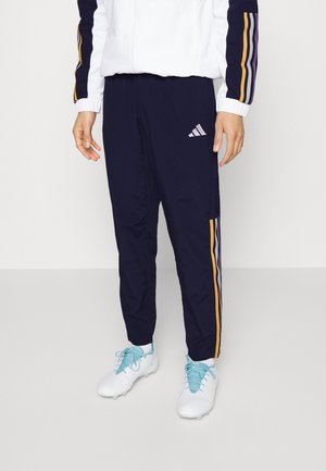 Pantalones de chándal azul marino con franjas laterales moradas y naranjas, combinados con zapatillas deportivas blancas y una chaqueta blanca con mangas a rayas a juego.