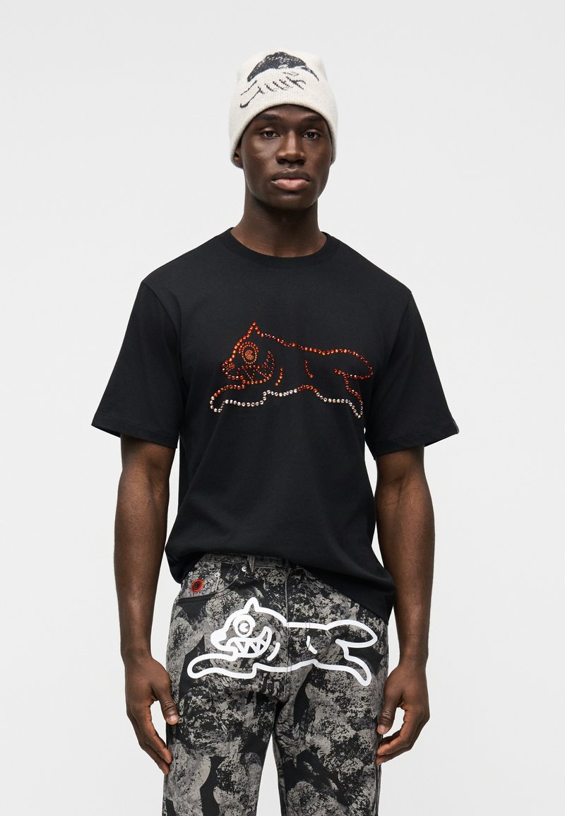 Μαύρο κοντομάνικο μπλουζάκι με πορτοκαλί και λευκό σχέδιο γάτας από στρας. Γκρι τζιν με σχέδια που διαθέτουν ένα λευκό γραφικό γάτας στην πίσω τσέπη.